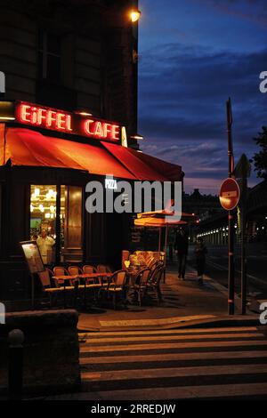 Eiffel Cafe Spätsommerabend, rotes Neonschild leuchtet vor dem blauen Nachthimmel, Café-Stühle, Straßenlaternen auf Grenelle und Pont de Bir-Hakeim Stockfoto