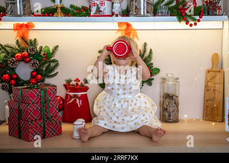 Weihnachtsküchenmädchen. Porträt eines charmanten kleinen Mädchens im Alter von 2 Jahren, sie sitzt auf einem Tisch in einem weißen Kleid und trinkt aus einem roten Becher in einem dekorierten Stockfoto