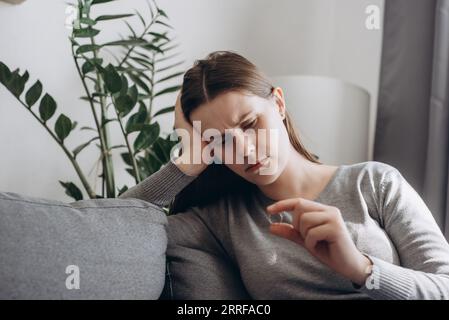 Frustrierte unglückliche verzweifelte Frau sitzt auf dem Sofa und hält den Verlobungsring in der Hand, gestresste junge Frauen leiden unter Beziehungsabbruch oder Verlobte Wette Stockfoto