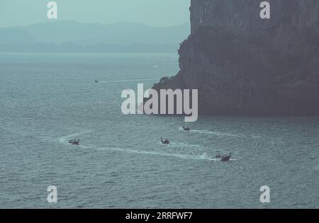 Langboote fahren um einen Felsvorsprung eines Kalksteingebirges und segeln auf dem abendlichen Meer. Dies ist die Route von Ao Nang Town nach Railay Beach Stockfoto