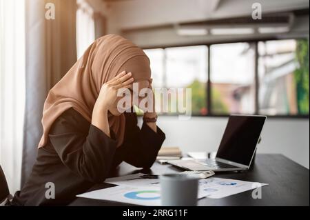 Eine gestresste und nachdenkliche, tausendjährige asiatische, muslimische Geschäftsfrau in einem Hijab liest und untersucht Geschäftsberichte mit einem ernsten Gesicht, besorgt über sie Stockfoto