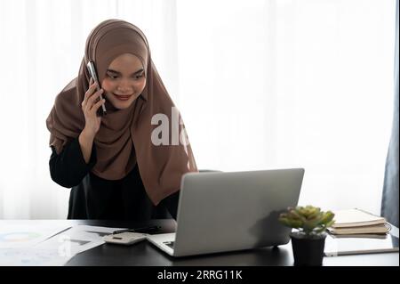 Eine professionelle und selbstbewusste asiatische muslimische Geschäftsfrau oder eine weibliche Finanzassistentin spricht mit ihrem Kunden am Telefon, während sie sich den Bettler anschaut Stockfoto