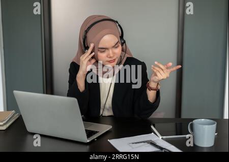 Eine verwirrte und nachdenkliche asiatisch-muslimische Callcenter-Betreiberin mit Headset und Hijab löst Kundenprobleme während des Anrufs, Konsul Stockfoto