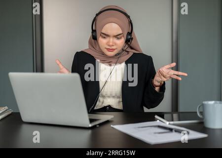 Eine gestresste und ernsthafte asiatische muslimische Kundendienstmitarbeiterin oder technische Supportmitarbeiterin, die ein Headset und einen Hijab trägt, unterhält sich stressig Stockfoto