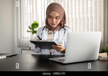 Eine professionelle und schöne asiatische muslimische Ärztin in Uniform und Hijab konzentriert sich darauf, ihre medizinischen Fallinformationen auf einem Klemmbrett, wo, zu überprüfen Stockfoto