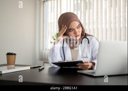 Eine gestresste und ernsthafte asiatische muslimische Ärztin in Uniform und Hijab schaut mit einem nachdenklichen Gesicht aus dem Fenster, während sie an ihrem Schreibtisch sitzt Stockfoto