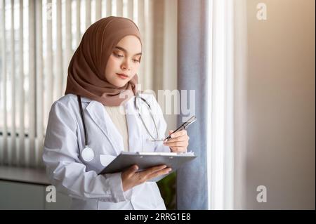 Eine fokussierte und professionelle asiatische muslimische Ärztin in Uniform und Hijab überprüft ihre medizinischen Fälle auf einem medizinischen Klemmbrett, während sie bei Th steht Stockfoto