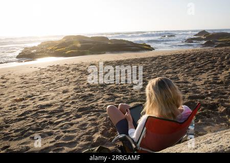 Reife Frau benutzt Tablet, während sie im Strandstuhl am Meer sitzt Stockfoto