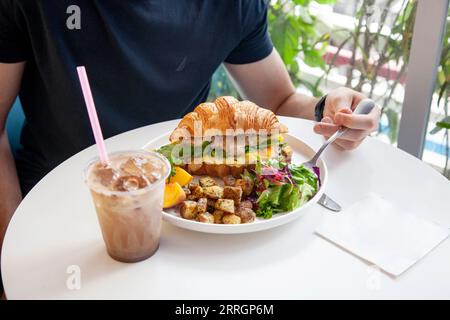 Mann, der ein köstliches Frühstück mit Croissant-Sandwich und Eiskaffee hat. Stockfoto