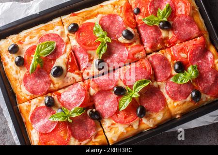 Frisch gebackene rechteckige Pizza mit Käse, Salami, Oliven und Tomaten aus nächster Nähe in einem Backblech auf dem Tisch. Horizontale Draufsicht von oben Stockfoto