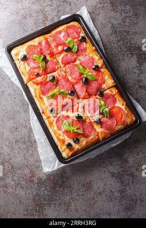 Rechteckige Pizza mit Käse, Wurst, Oliven und Tomaten in einem Backblech auf dem Tisch. Vertikale Draufsicht von oben Stockfoto