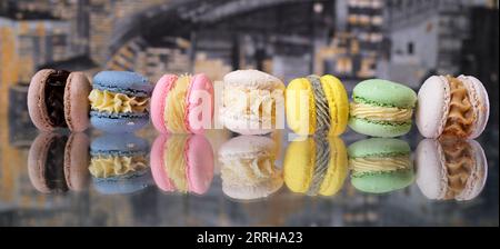 Mehrfarbige Macarons liegen auf einem Glastisch mit Reflexion vor dem Hintergrund einer nächtlichen Stadttapete rosa grün gelb beige blaue Schokoladen-Macarons. Köstliches süßes Dessert Stockfoto