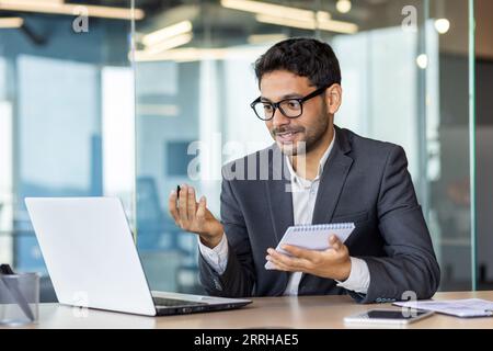 Junger erfolgreicher hispanischer Mann, der sich Online-Video ansieht kur Webinar, Geschäftsmann, der am Arbeitsplatz im Büro studiert, Daten in Notizbücher schreibt, Mann, der die Qualifizierung verbessert. Stockfoto