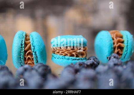 Heidelbeer-Macarons Beeren verstreut auf dem Tisch in der Nähe von köstlichen Süßigkeiten Karamell Füllung von blauen Macarons verschwommene Fokusfläche für Textwerbung Postkarte Party Einladung. Köstliches süßes Dessert Stockfoto