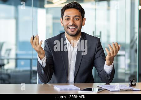 Videoanruf per Webcam ansehen, junger arabischer Mann im Büro, der mit Kollegen aus der Ferne spricht, Mann, der die Kamera ansieht, lächelt am Arbeitsplatz im Büro. Stockfoto