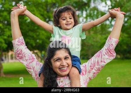 Glückliche junge indische Mutter, die ihre Tochter im Sommerpark auf den Schultern trägt. Familien- und Mutterschaftskonzept. Stockfoto