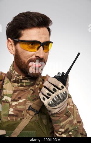 Gut aussehender Militärmann in gelben taktischen Gläsern, der mit Walkie-Talkie spricht. Porträt eines lächelnden Soldaten in Tarnuniform, mit tragbarem Radio, Berichterstattung, isoliert auf weiß. Verbindungskonzept. Stockfoto