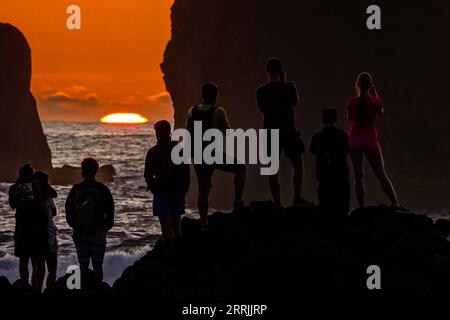 Junge Leute treffen sich, um die riesigen Meeresstapel zu beobachten, die vom Sonnenuntergang auf Ponta dos Mosteiros in Sao Miguel, Azoren, Portugal, umrahmt werden. Stockfoto