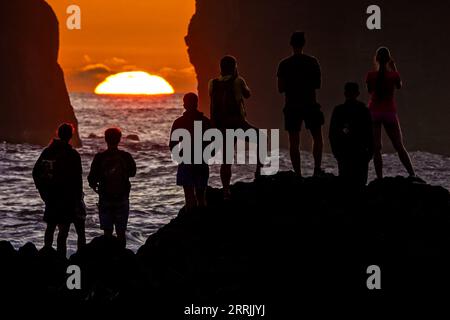 Junge Leute treffen sich, um die riesigen Meeresstapel zu beobachten, die vom Sonnenuntergang auf Ponta dos Mosteiros in Sao Miguel, Azoren, Portugal, umrahmt werden. Stockfoto