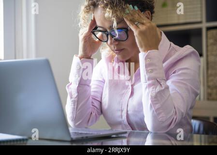 Schmerzhafte Kopfschmerzen bei erwachsener Frau am Arbeitsplatz, die den Tempel berührt. Nachdenkliche, moderne Online-Mitarbeiter, Frauen vor einem Laptop an der Rezeption. Geschäftliche Probleme und Dame besorgt. Überarbeit unzufriedene Freiberuflerin Stockfoto