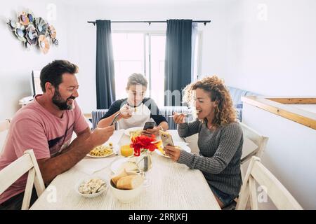 Glückliche junge Familie genießen gemeinsam zu Hause ein Mittagessen mit dem Mobiltelefon. Süchtig machende Online-Mobiltelefone in sozialen Medien, die im Wohnzimmer essen und sich lustige Displays anschauen. Konzept des SuchtSmartphones Stockfoto