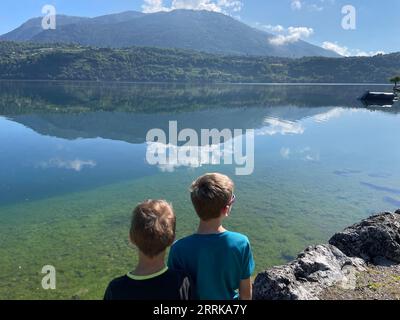 Zwei Jungen, die den ruhigen Caldonazzo-See im Trentino, den Caldonazzo-See, das Suganertal, Valsugana, den Levico-See, Lago di Levico, Natur, Wasser, Berge, Aktivität, Sonne, Wolken, Caldonazzo, Trentino Südtirol, Italien Stockfoto
