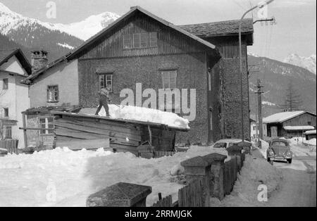 Tirol, Österreich. c.1960 – St. Anton am Arlberg. Ein kleiner Junge steht auf dem Dach eines rustikalen hölzernen Nebengebäudes und entfernt mit einem Spaten eine dicke Schicht Schnee von seinem Dach. Dahinter befindet sich ein Haus im traditionellen Stil mit Verkleidung an den Wänden. Ein Volkswagen Beetle Auto steht auf der angrenzenden Straße. Schneebedeckte Berge der Ostalpen sind in der Ferne sichtbar. Stockfoto