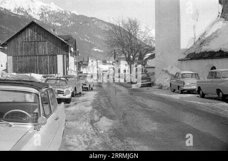 Tirol, Österreich. c.1960 – Blick auf die Dorfstraße im Dorf St. Anton am Arlberg. Auf der rechten Seite befindet sich St. Anthony von Padua Kirche. Die Autos werden auf beiden Seiten der Straße geparkt, die sich in einem schlampigen Zustand befindet. Hotels und die schneebedeckten Berge der Ostalpen sind in der Ferne sichtbar. Stockfoto