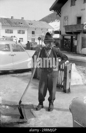 Tirol, Österreich. c.1960 – St. Anton am Arlberg. Ein älterer Mann mit einem buschigen Hammelbart und einem traditionellen Tiroler Hut steht mit einer langgezogenen Schaufel, mit der er Schnee vom Pflaster entfernt. Hotels und die schneebedeckten Berge der Ostalpen sind in der Ferne sichtbar. Stockfoto