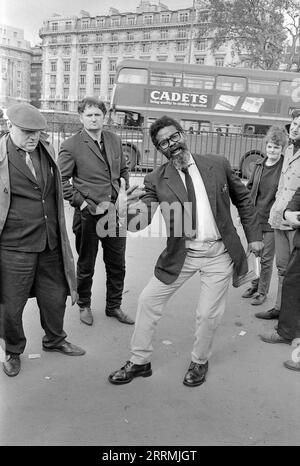 London. 1965: Ein Evangelist predigt in Speaker’s Corner, Hyde Park an der Kreuzung von Cumberland Gate, in der Nähe von Marble Arch, London. Der Prediger hält eine Bibel in der einen Hand und gestikuliert mit der anderen. Eine Gruppe von Zuschauern hat sich um ihn herum versammelt. Das Gebäude an der 140 Park Lane ist in der Ferne sichtbar. Ein Doppeldeckerbus in London fährt mit einer Werbung für Cadets Cigarettes vorbei. Stockfoto