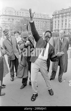 London. 1965: Ein Evangelist predigt in Speaker’s Corner, Hyde Park an der Kreuzung von Cumberland Gate, in der Nähe von Marble Arch, London. Der Prediger hält eine Bibel in der einen Hand und hebt seine andere Hand in die Luft. Eine Gruppe von Zuschauern hat sich um ihn herum versammelt. Ein älterer Mann, der einen Regenmantel und eine flache Kappe trägt und seine Hand in der Faust zusammenballt und den Prediger verspottet. Das Cumberland Hotel und das Gebäude an der 140 Park Lane sind in der Ferne zu sehen. Stockfoto