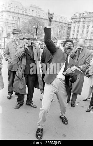 London. 1965: Ein Evangelist predigt in Speaker’s Corner, Hyde Park an der Kreuzung von Cumberland Gate, in der Nähe von Marble Arch, London. Der Prediger hält eine Bibel in der einen Hand und zeigt seine andere Hand zum Himmel. Eine Gruppe von Zuschauern hat sich um ihn herum versammelt. Ein älterer Mann, der einen Regenmantel und eine flache Kappe trägt, verspottet den Prediger, indem er ihm eine Himbeere durch die Hand bläst. Das Cumberland Hotel und das Gebäude an der 140 Park Lane sind in der Ferne zu sehen. Stockfoto