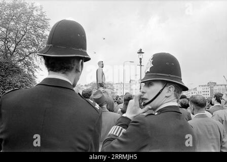 London. 1965: Ein Mann, der auf einer Plattform steht und den Panafrikanischen Nationalkreuzzug vertritt, spricht in der Speaker’s Corner im Hyde Park, London, vor einer Menge von Zuschauern. Zwei Metropoliten stehen im Vordergrund, einer schaut über seine Schulter, das Pflichtarmband auf seinem Ärmel ist zu sehen. Stockfoto