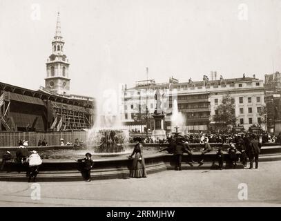 London, England. 1911: Blick auf die Springbrunnen am Trafalgar Square, London. Das Foto zeigt die originalen Springbrunnen-Mittelstücke aus den 1840er Jahren Diese wurden nach dem Zweiten Weltkrieg durch einen neuen Entwurf von Sir Edwin Lutyens ersetzt. Eine provisorische Tribüne, die links zu sehen ist, und ein Balkon, der an der Vorderseite des Morley’s Hotel gegenüber angebracht ist, wurden beide zur Vorbereitung der Krönung von König Georg V. im Juni 1911 errichtet. Zwischen den Springbrunnen befindet sich die Statue von General Gordon, die 1943 entfernt und verlegt wurde. Die St. Martin-in-the-Fields-Kirche ist in der Ferne zu sehen. Stockfoto