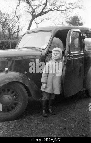 England. c.1960: Ein kleiner Junge mit Mantel, gummistiefeln und Wollmütze steht vor der offenen Tür eines Standard-Acht-Limousine-Wagens aus dem Jahr 1948. Stockfoto