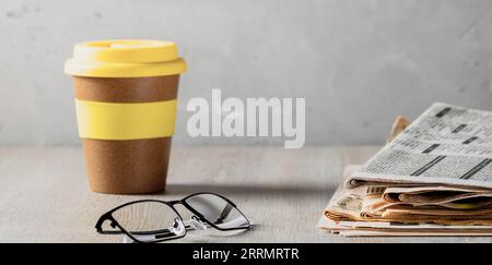 Ich lese die Morgennachrichten. Ein Stapel Zeitungen, Gläser und ein Glas Kaffee auf einem Holztisch, der von der Morgensonne beleuchtet wird. Stockfoto