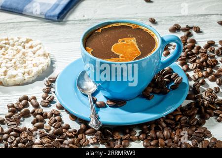 Morgenkaffee. Eine blaue Tasse Kaffee mit Schaum auf einem Holztisch, umgeben von verstreuten Kaffeebohnen. Im Hintergrund gibt es einen Reiskuchen und eine Co Stockfoto