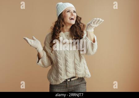 Hallo Winter. Lächelnde, stylische Frau in beigem Pullover, Fäustlingen und Hut isoliert auf beigem Hintergrund, um Schnee zu fangen. Stockfoto