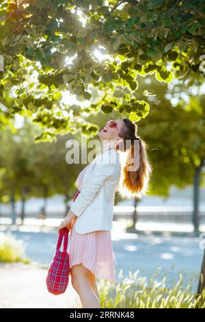 Glückliche, stilvolle Frau in rosa Kleid und weißer Jacke in der Stadt mit roter Tasche und Sonnenbrille. Stockfoto