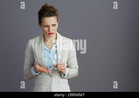 Elegante 40-jährige Arbeiterin im leichten Business-Anzug mit Glas Wasser und Pille vor grauem Hintergrund. Stockfoto