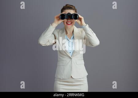 Glückliche trendige mittelalte Geschäftsinhaberin Frau in einem leichten Business-Anzug mit Fernglas isoliert auf grauem Hintergrund. Stockfoto