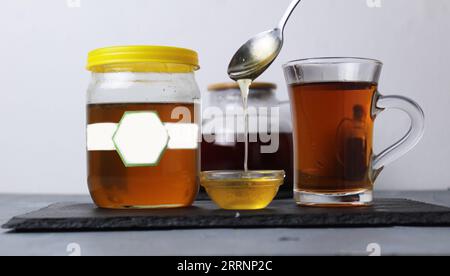 Stillleben mit Honig gießt Honig in eine Tasse, fließt in einem dünnen Strom, daneben ein Glas Tee, ein Glas Honig auf grauem Hintergrund. Behandlungswit Stockfoto