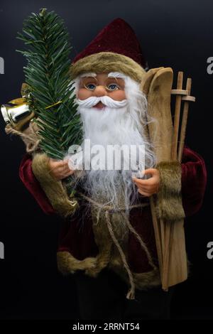 Weihnachtsmann-Spielzeug mit Holzskiern in der einen Hand und einem Tannenzweig mit einer Glocke in der anderen auf schwarzem Hintergrund Stockfoto