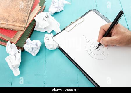 Männliche Hand, die ein Ziel auf einem weißen Klemmbrett mit einem schwarzen Stift neben Vintage-Büchern und zerknittertem Papier zeichnet Stockfoto