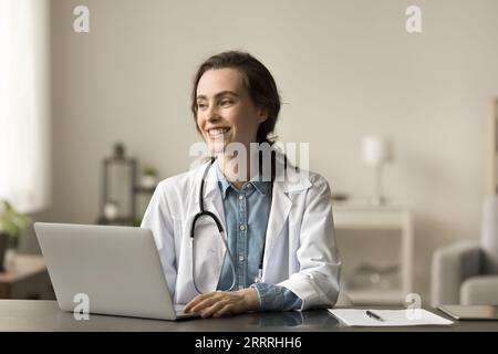 Fröhliche junge Ärztin im weißen Mantel am Tisch sitzend Stockfoto