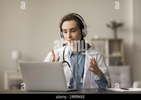 Seriöser Arzt im weißen Mantel und Kopfhörer, der am Laptop spricht Stockfoto