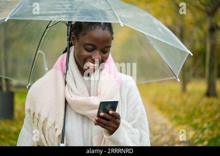 Afrikanische Frau mit einem Schirm, die lächelt und im Herbst telefoniert. Stockfoto