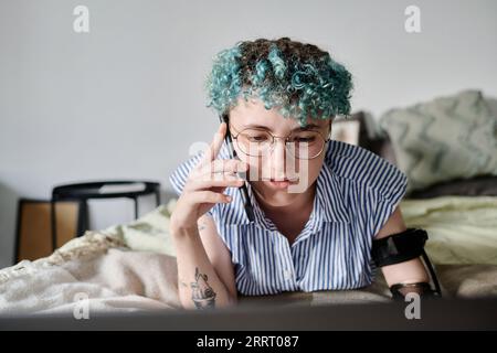 Junge Frau mit gefärbten lockigen Haaren, die auf dem Handy telefoniert, während sie einen Laptop benutzt Stockfoto