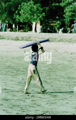 Kleiner Junge, der ein Surfbrett am Strand auf Bali trägt Stockfoto