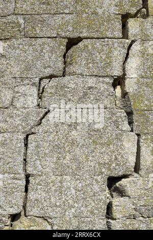 Eine alte, mit Moos bedeckte Steinmauer mit tiefen Rissen und eingestürzten Steinen. Große diagonale Risse an der alten Wand. Leerzeichen kopieren. Selektiver Fokus. Stockfoto